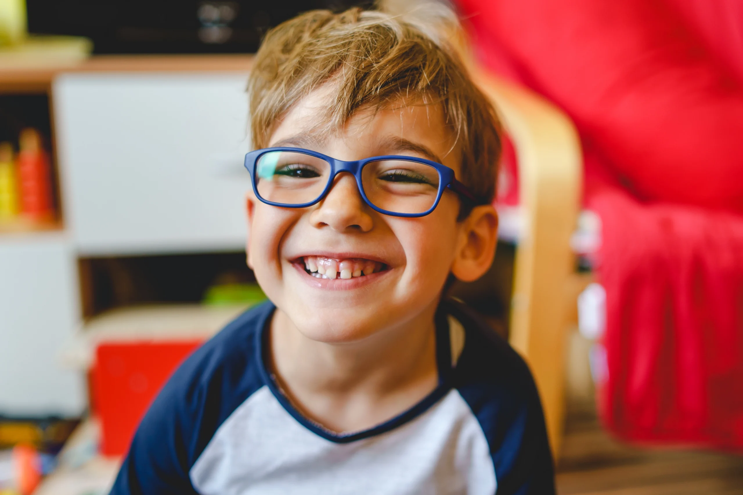 A little boy in blue framed glasses grins at the camera.