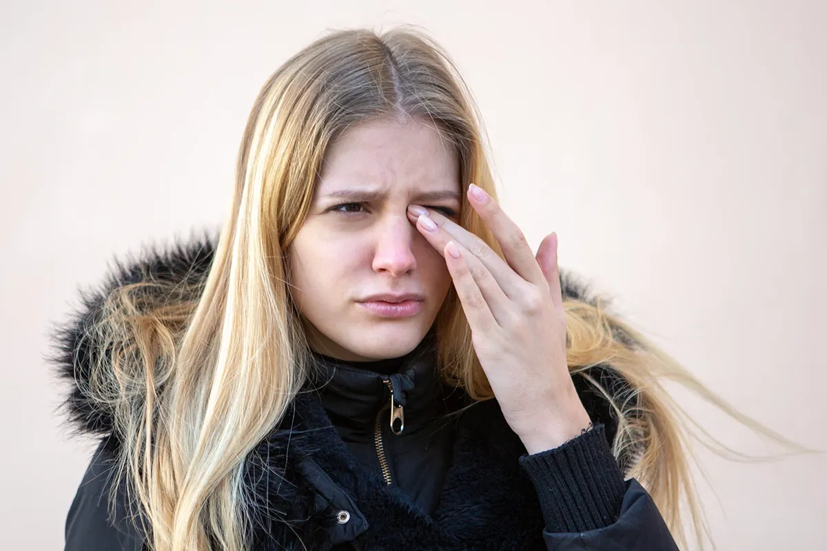 A young woman in a winter coat rubs her eyes in discomfort.